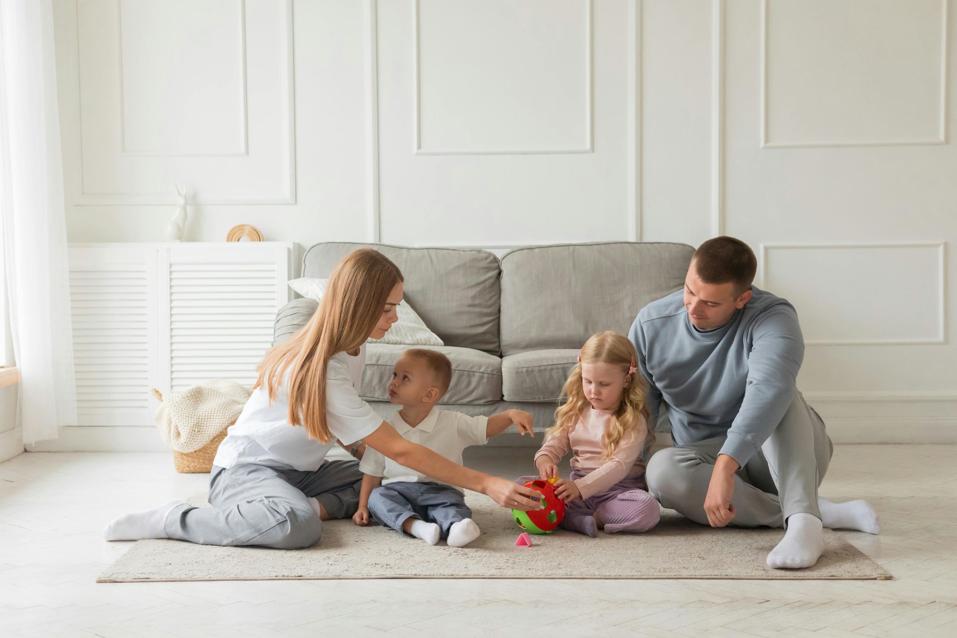 A family of four, including a mother, father, a young boy, and a young girl, are sitting together on a rug on the floor of a brightly lit, modern living room. They are gathered around a red and green shape-sorter toy, with the mother guiding the children as they play in front of a gray sofa.