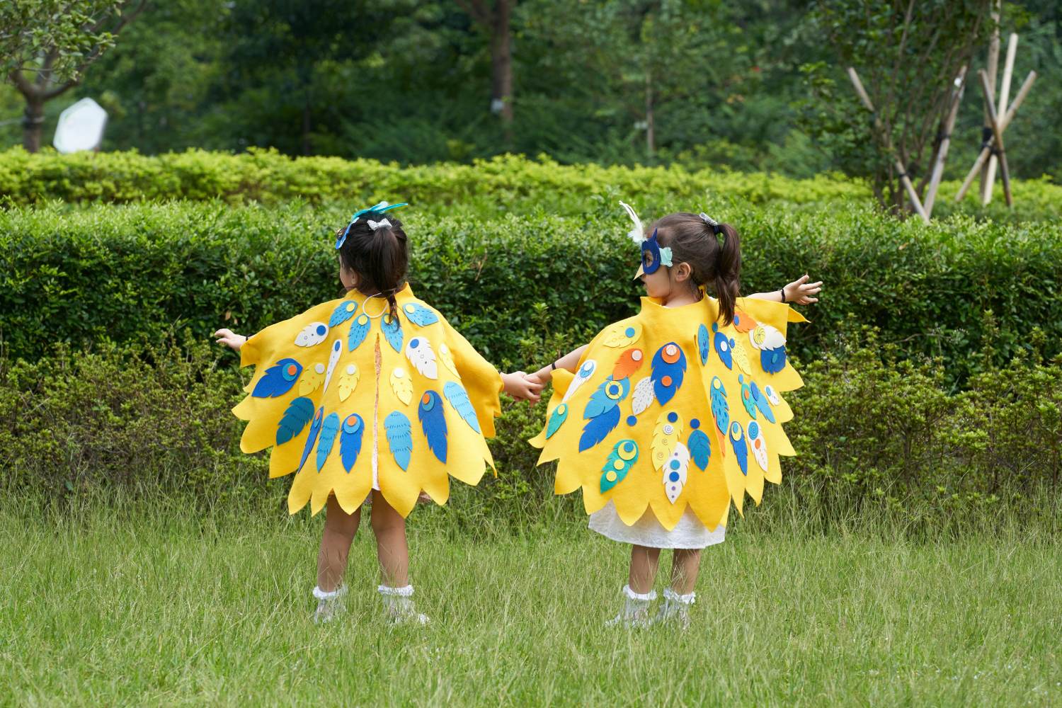 Two young girls, seen from behind, are standing hand-in-hand in a grassy park. They are wearing matching homemade yellow capes designed to look like bird wings, decorated with colorful felt feathers in shades of blue, white, and orange. One girl wears a blue mask over her eyes. They have their arms outstretched, pretending to fly in front of a lush green hedge.