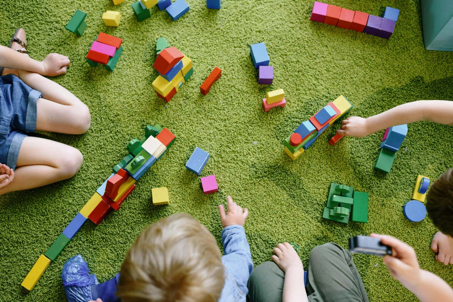 An overhead view shows a group of children sitting on a green carpet, playing with colorful wooden building blocks. The blocks are scattered across the floor, and the children are building various structures.