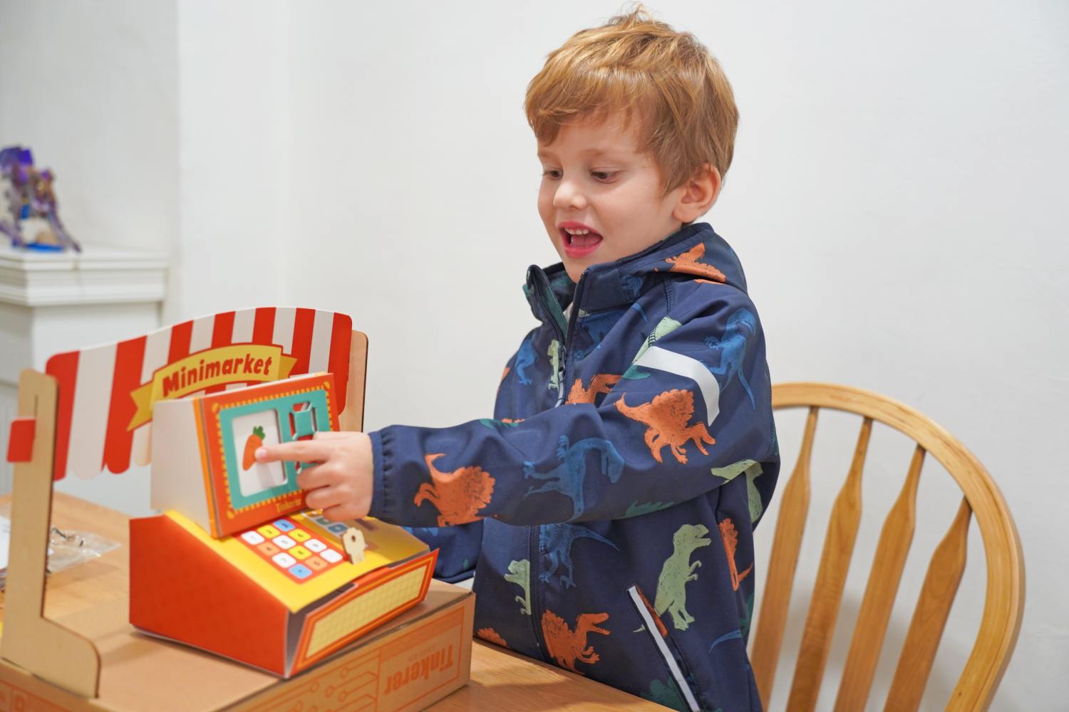 A young boy with reddish-blond hair, wearing a blue jacket with a dinosaur pattern, sits at a wooden table and excitedly plays with a colorful cardboard toy cash register. The toy is designed to look like a minimarket stall with a red and white striped awning.