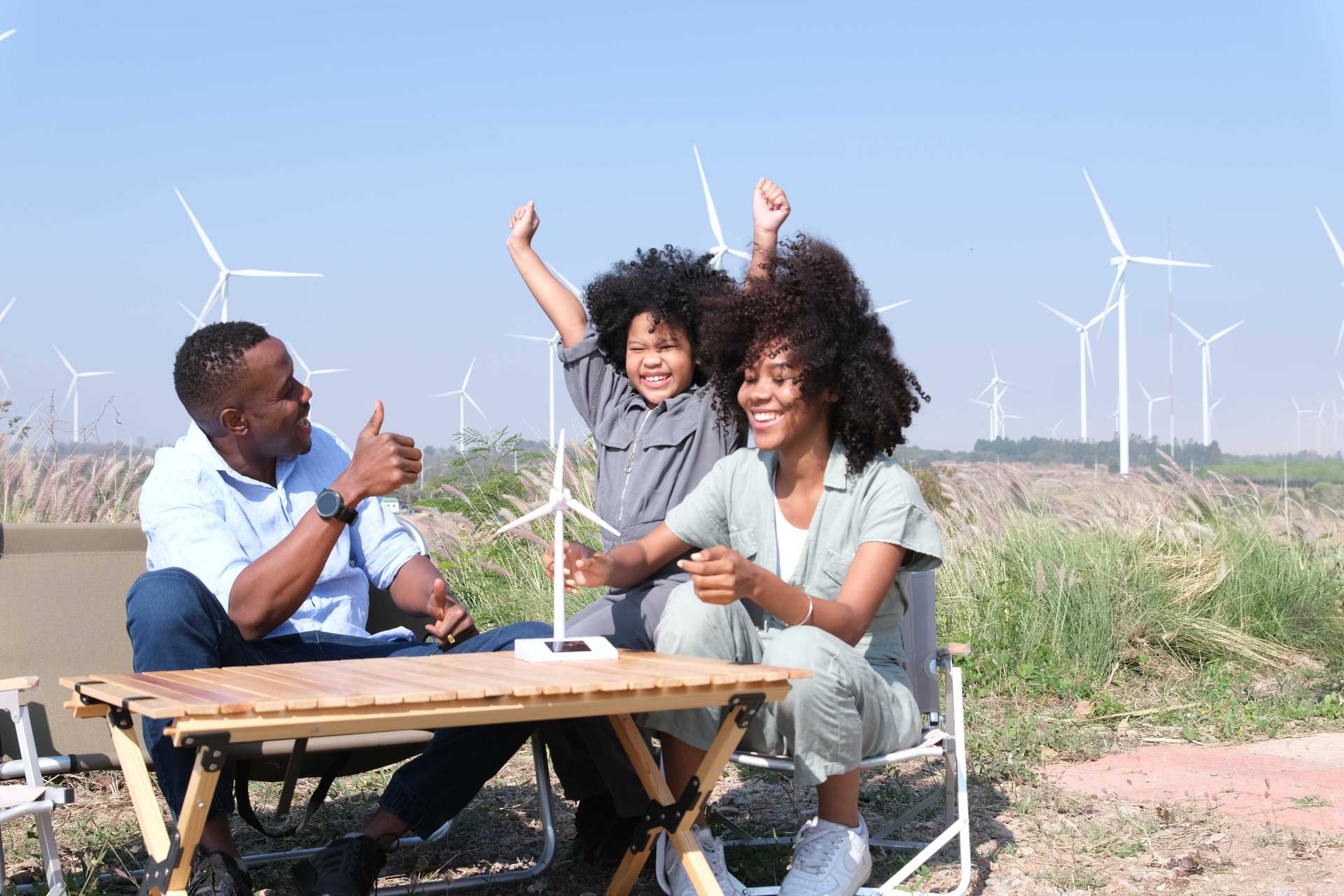 A joyful family enjoys a picnic in a field of tall grass, with spinning wind turbines dotting the horizon. The father sits in a folding chair, giving a thumbs-up, while the mother and son share a wooden folding table. The boy proudly holds up a small model wind turbine, his face beaming with excitement. This heartwarming scene captures the essence of family bonding, environmental awareness, and hands-on learning. The real turbines in the background mirror the family's enthusiasm for clean energy, creating a powerful image of hope and commitment to a sustainable future. Their shared smiles and engaged postures reflect not just a pleasant day out, but a family united in their passion for making a positive difference in the world.