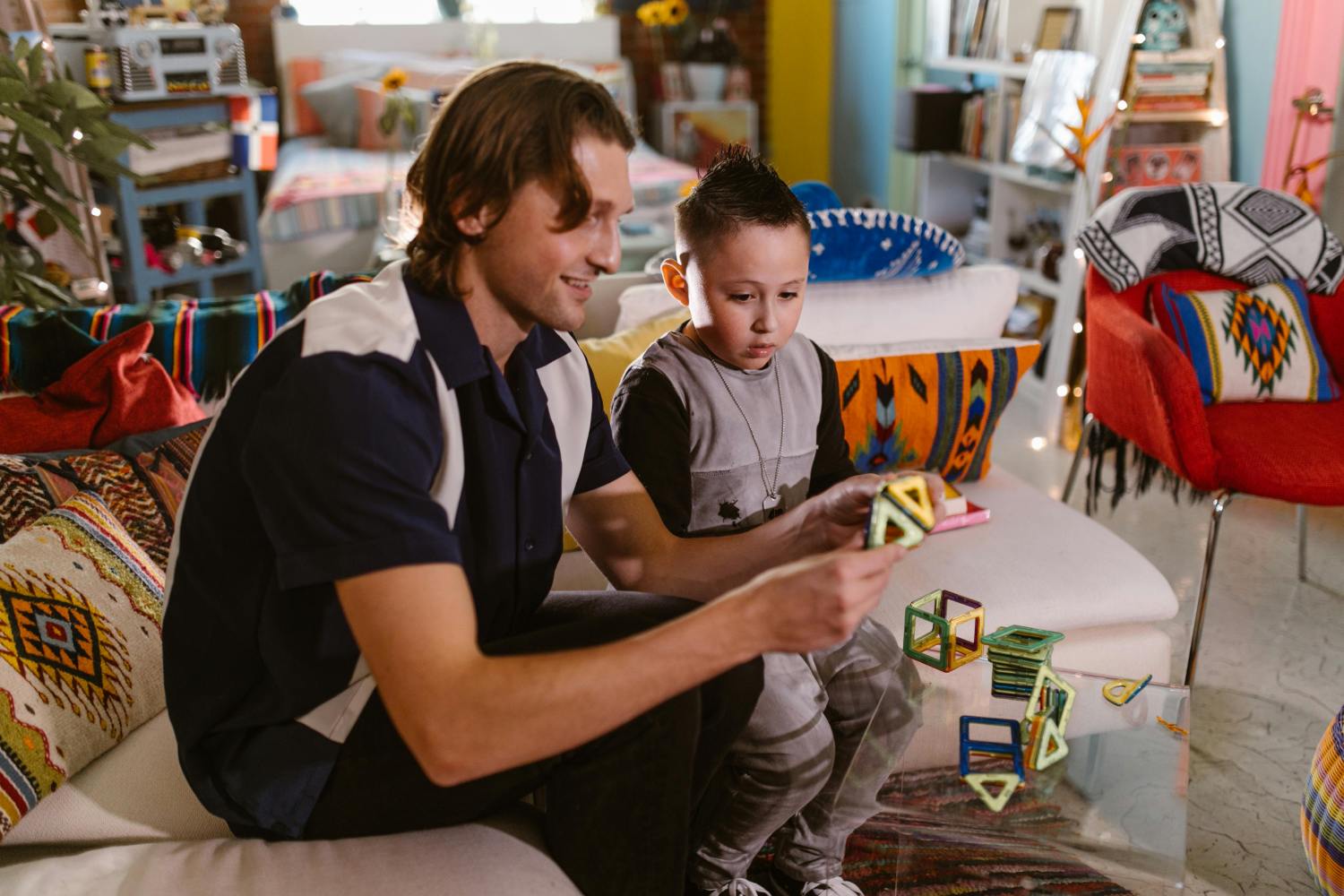 A man with shoulder-length brown hair and a young boy with a mohawk haircut are sitting together on a white couch in a colorful, eclectically decorated living room. They are both focused on playing with colorful, magnetic geometric building tiles, creating a structure on a clear coffee table in front of them. The man is smiling as he interacts with the boy.