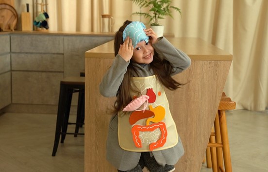 A young girl wearing an apron with a colorful human organ illustration from a STEM project, playfully holding a blue fabric eye mask over her head, looking at the camera with a mischievous smile.