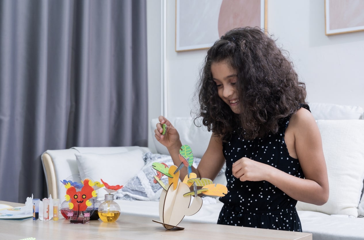 A young girl with long, curly hair is sitting at a table, happily assembling a wooden toy plant from a STEM box kit. She is holding a small green leaf piece in her hand, and her smile suggests she is enjoying the process. The table is covered in colorful craft supplies, including bottles of liquid, suggesting a science project. 