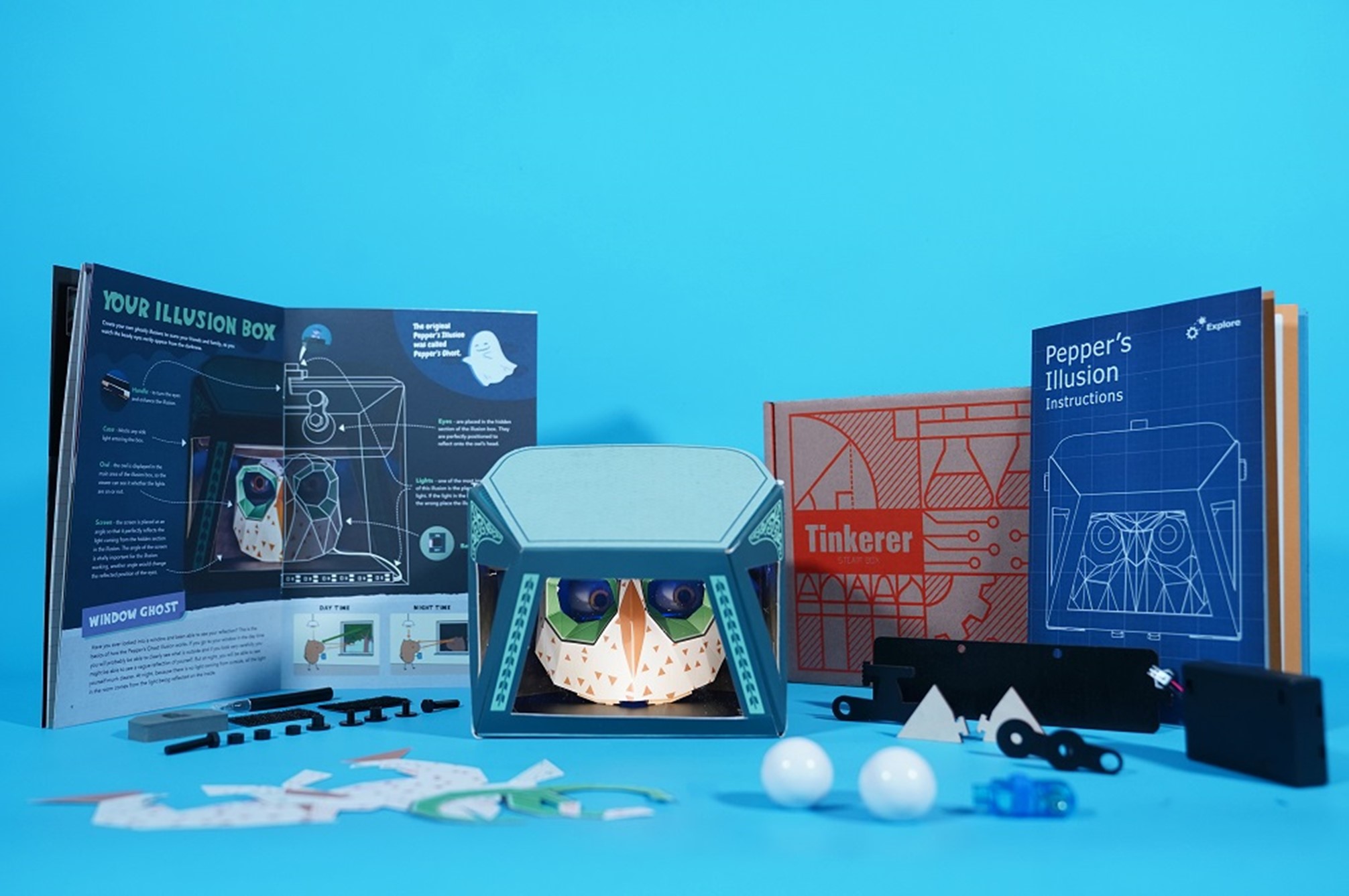 A Tinkerers STEM box featuring a Pepper's Illusion kit. The box includes a partially assembled cardboard owl illusion box, an instruction booklet, and various other components, including a small LED light, a few white balls, and a blue plastic piece. The kit is laid out on a bright blue background, suggesting a fun and engaging project.