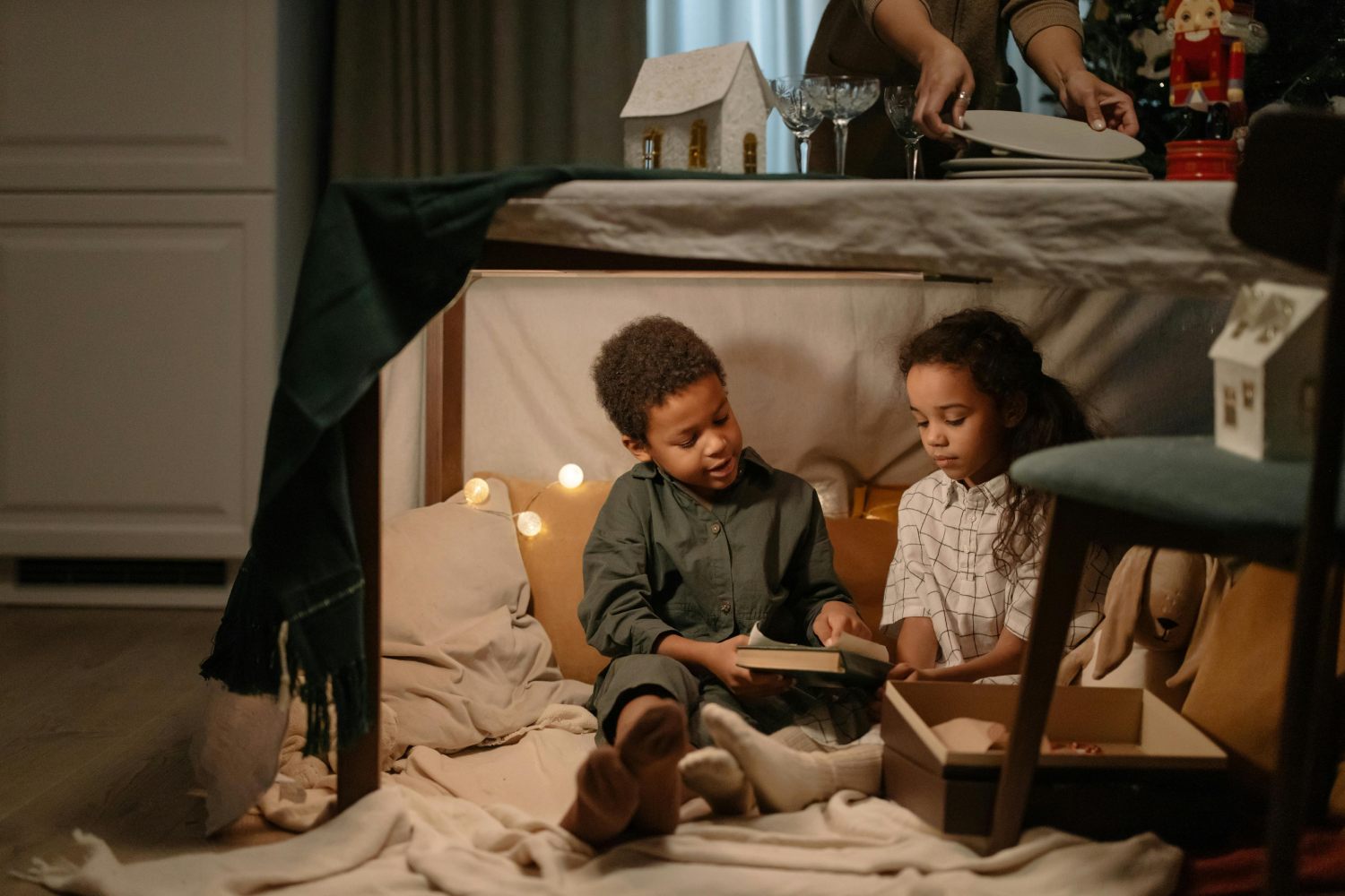 Two young children, a boy and a girl, are sitting together inside a cozy blanket fort built under a table, looking at a book. The fort is lit by warm string lights. Above them, an adult's hands are seen setting the dining table for a meal, with holiday decorations visible.