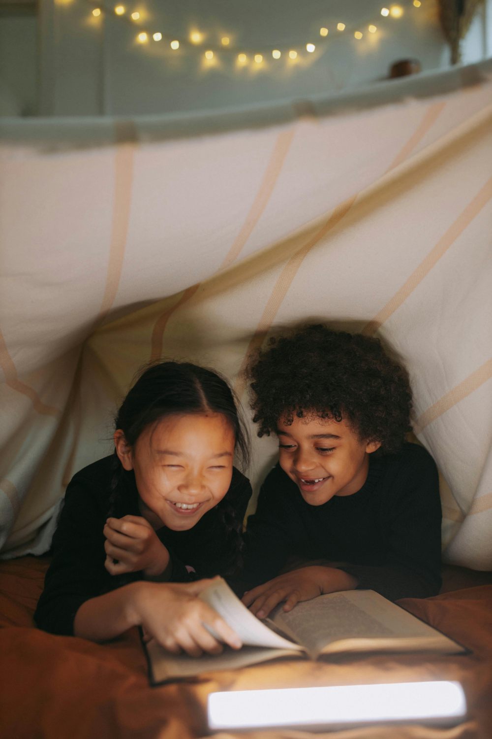 Two happy children, a girl with braided hair and a boy with curly hair, are lying inside a blanket fort, laughing as they read a book together. Their faces are illuminated by a light placed in front of the book, and string lights are visible in the blurred background.