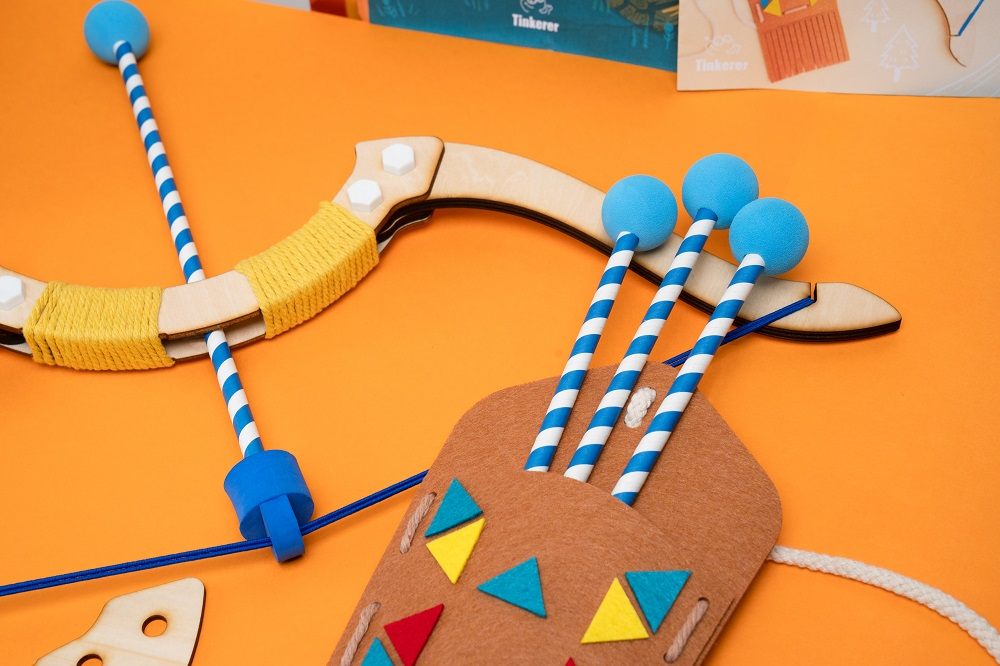 A close-up of a children's toy bow and arrow set on a bright orange background. The wooden bow has a yellow yarn-wrapped handle, and an arrow with a blue foam tip and a blue-and-white striped shaft is nocked on the string. A brown felt quiver, decorated with colorful triangles, holds several more arrows.