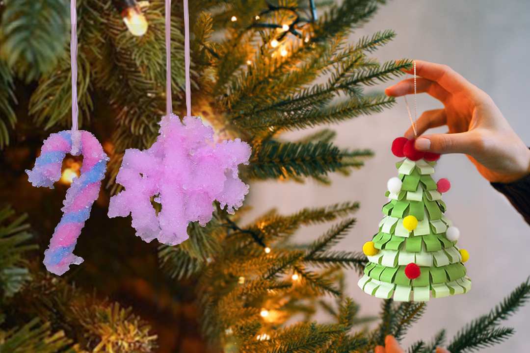 Three handmade Christmas ornaments are shown against the backdrop of a lit Christmas tree. Two of the ornaments appear to be crystallised, one shaped like a candy cane and the other like a snowflake. A hand holds a third ornament, a small Christmas tree crafted from folded green paper strips and decorated with small, colourful pom-poms.