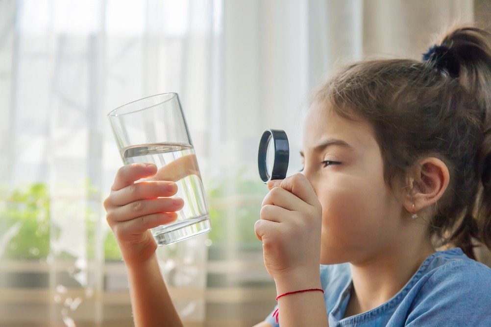 A young girl, full of curiosity, studies a glass of water using a magnifying glass. Her brow is knitted in concentration as she peers through the lens, uncovering the mysteries hidden in the seemingly mundane liquid. This moment captures the essence of observation and the thrill of discovery, highlighting a child's natural wonder about the world.