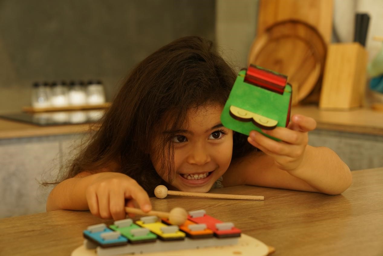 A child with long dark hair, leans over a wooden table, her eyes sparkling with joy as she plays with a wooden xylophone and a toy during a homeschool STEAM activity. She is holding a small green toy in her right hand and a wooden mallet in her left hand. The girl's smile is contagious, radiating pure happiness and the joy of discovery.