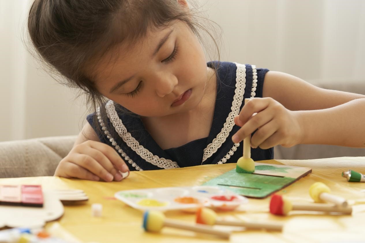 A young girl, with a focused expression, carefully paints a wooden craft project. Her hands are steady as she applies the paint, her brow furrowed in concentration. The scene is a testament to the power of creativity and the joy of bringing ideas to life.