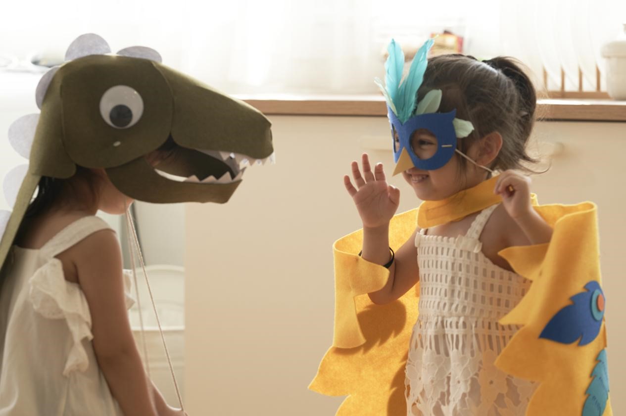 Two young girls, dressed in homemade costumes, are engaged in a playful interaction. One girl is wearing a dinosaur mask and the other a bird mask. They are both smiling and appear to be having a lot of fun. The scene is full of warmth and joy, suggesting a playful and loving relationship between the two girls.