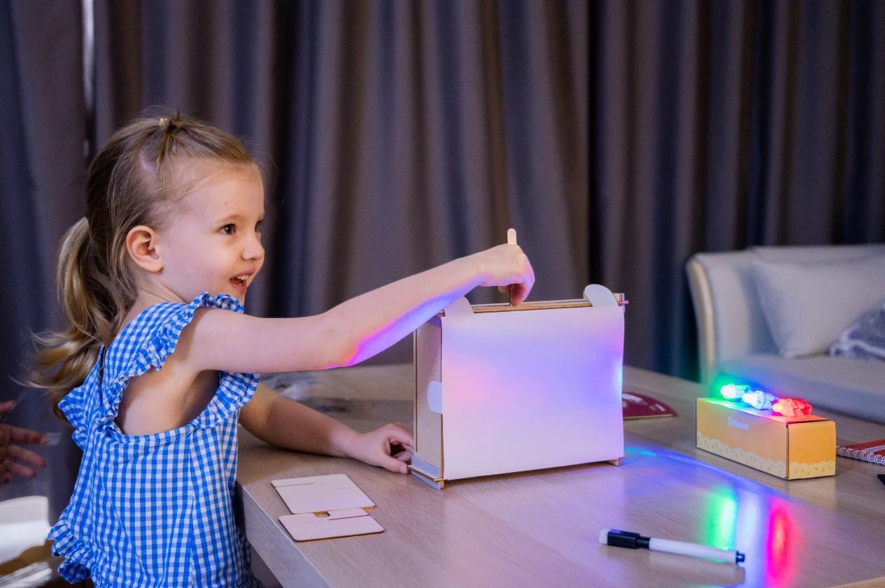 A childl, dressed in a blue and white gingham dress, is engrossed in creating a light show with a DIY light box. She is using colored lights to create different patterns and effects on the white surface of the box. The scene is filled with a sense of wonder and imagination, suggesting a child's love for experimenting and creating.