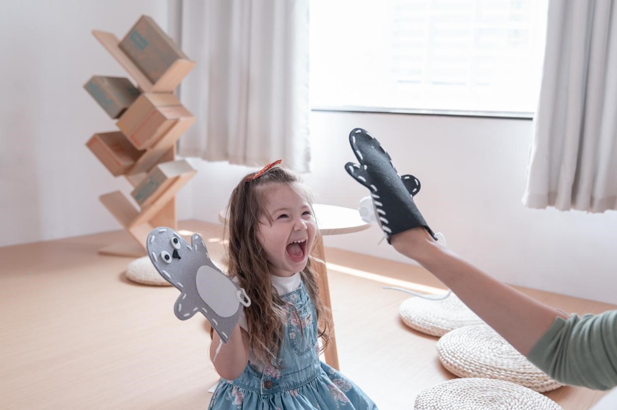 A little girl wearing a blue denim dress is laughing hysterically as she plays with a puppet. Her hand is inside a gray puppet. An adult hand is holding a black puppet shaped like a monster, and it appears to be high-fiving the girl. The scene is full of warmth and joy, suggesting a playful and loving relationship between the girl and the adult.