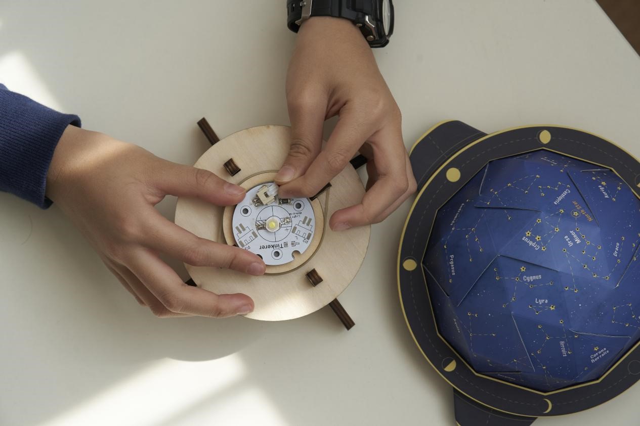 A person is assembling a DIY constellation projector. They are carefully connecting the LED light to the wooden base, while the constellation dome sits nearby. The image captures the process of creativity and innovation, as the person brings together different elements to create a unique and beautiful object.