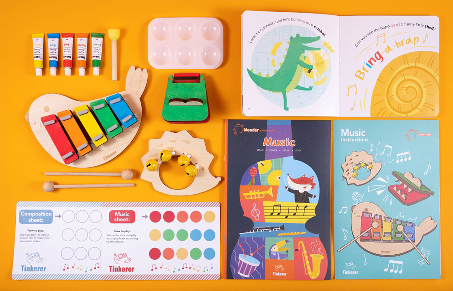 A flat lay arrangement of children's musical and art supplies is displayed on a vibrant orange background. The items include a bird-shaped xylophone, a wooden rattle with bells, small tubes of paint, a paint palette, wooden mallets, and several illustrated booklets or activity sheets related to music. The booklets feature cartoon animals playing instruments and instructions for musical activities. Two of the activity sheets appear to be guides for composing and playing music using colors and the xylophone. The scene suggests a fun and educational music-themed kit or activity set for children.