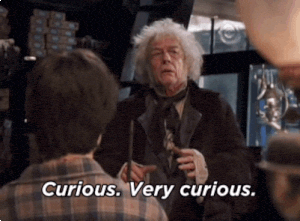 An elderly man with long white hair, wearing a dark suit and cravat, examines a wand. He holds the wand delicately in his hands, turning it over and inspecting it closely. He then looks up at the person who presumably presented the wand, a young boy with dark hair. The boy, facing the man, is only partially visible. The setting appears to be a shop, with shelves filled with various items blurred in the background. The man speaks, his words Curious. Very curious. appearing as text on the screen.
