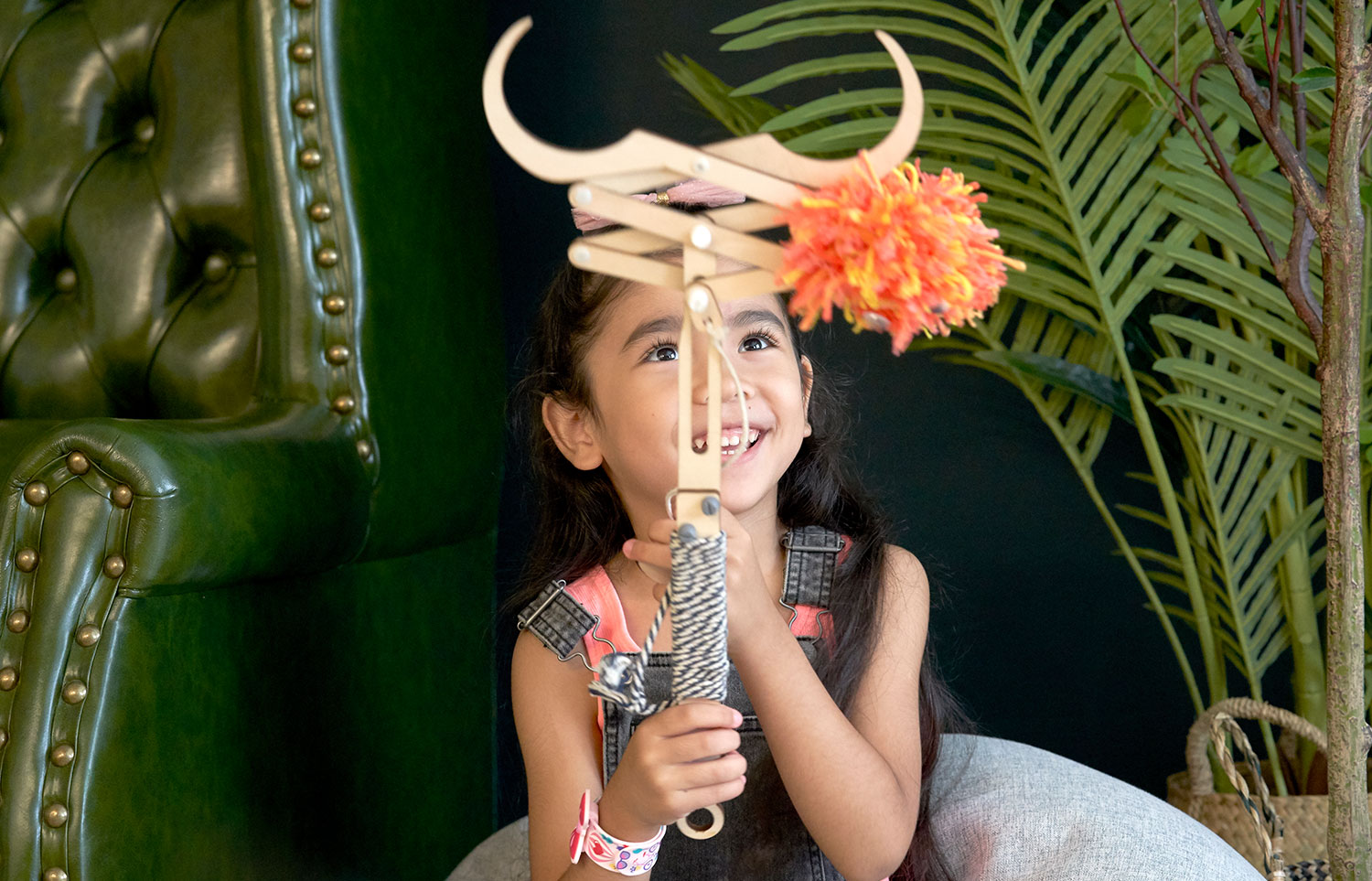 A young girl with long dark hair smiles as she looks up at a wooden toy she holds in her hands. The toy resembles a puppet or a small crane, with long, curved wooden arms and a bright orange and yellow pom-pom or tassel hanging from the center. She wears a coral pink top and a denim jumper. A plush, tufted dark green armchair is partially visible in the background to the left, and large green plants provide a backdrop to the right. The setting appears to be indoors, possibly a playroom or a child's bedroom.