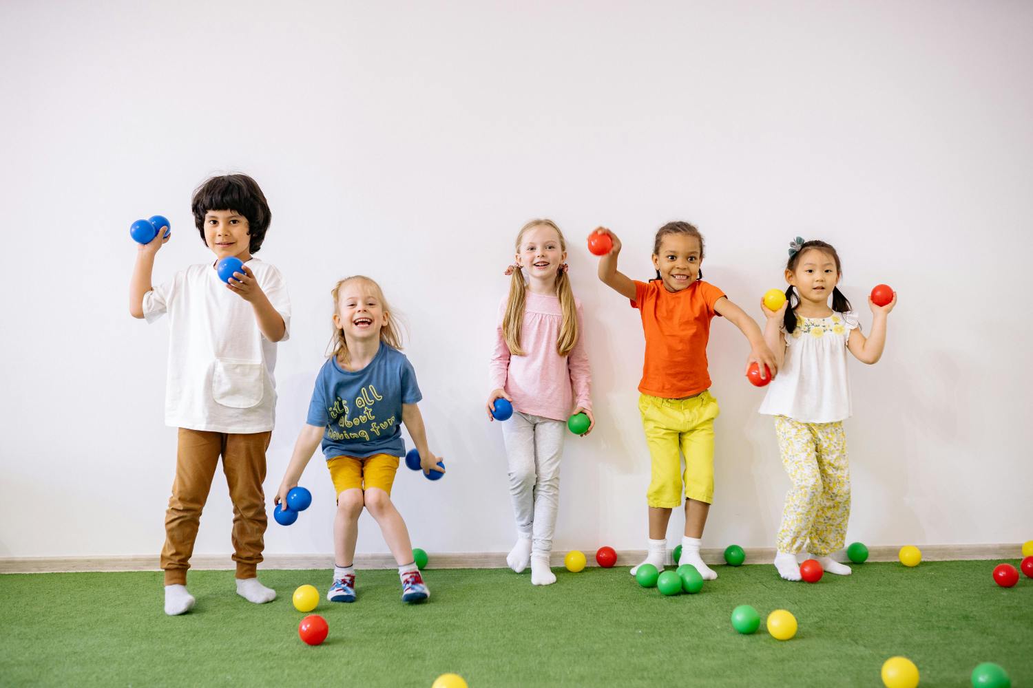 A group of five diverse and happy children are lined up against a white wall, standing on green artificial grass. They are all smiling and energetically posing while holding small, colorful balls, with more red, yellow, green, and blue balls scattered on the floor around their feet.