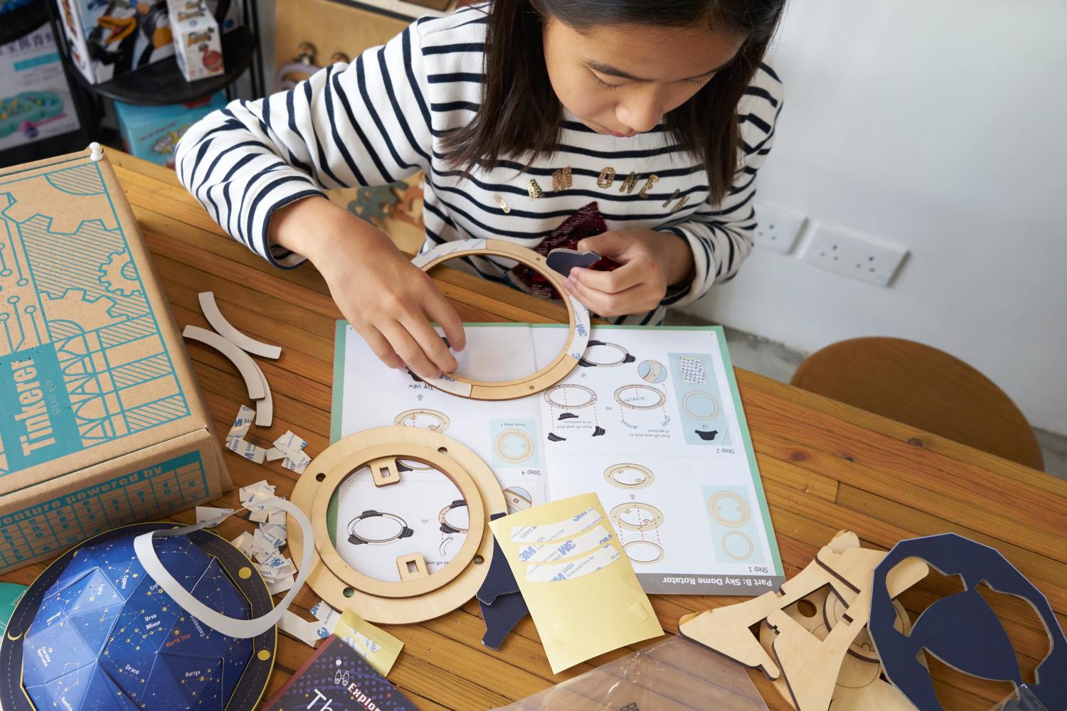 A young girl with dark hair, wearing a black-and-white striped shirt, sits at a wooden table and concentrates on assembling a model from a Tinkerer STEAM kit. The table is covered with various components of the kit, including wooden cutout pieces, an instruction manual for a Sky Dome Rotator, and parts of a celestial globe.