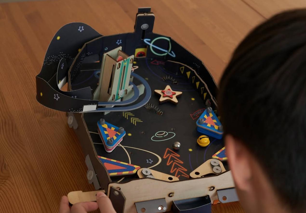 An over-the-shoulder view of a person playing with a DIY pinball machine made of cardboard and wood. The machine has a space theme, with a dark playfield decorated with colorful illustrations of planets, stars, and UFOs. A silver pinball is in the middle of the playfield, and the player's hands are visible at the bottom, operating the flipper controls.