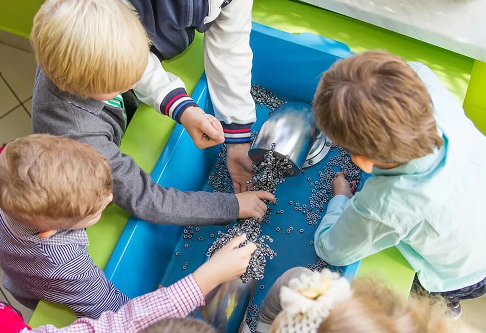 The photograph shows a high-angle, close-up view of several young children actively engaged in a hands-on activity. They are gathered around a bright blue plastic bin filled with numerous small, metallic washers or similar components. A metallic chute or funnel is positioned above the bin, from which more of the metallic pieces are flowing. The children appear to be carefully sorting or manipulating the components, their hands reaching into the bin. They are dressed in a variety of casual clothing. The background is partially visible and suggests an indoor setting. The overall impression is one of playful learning and exploration.