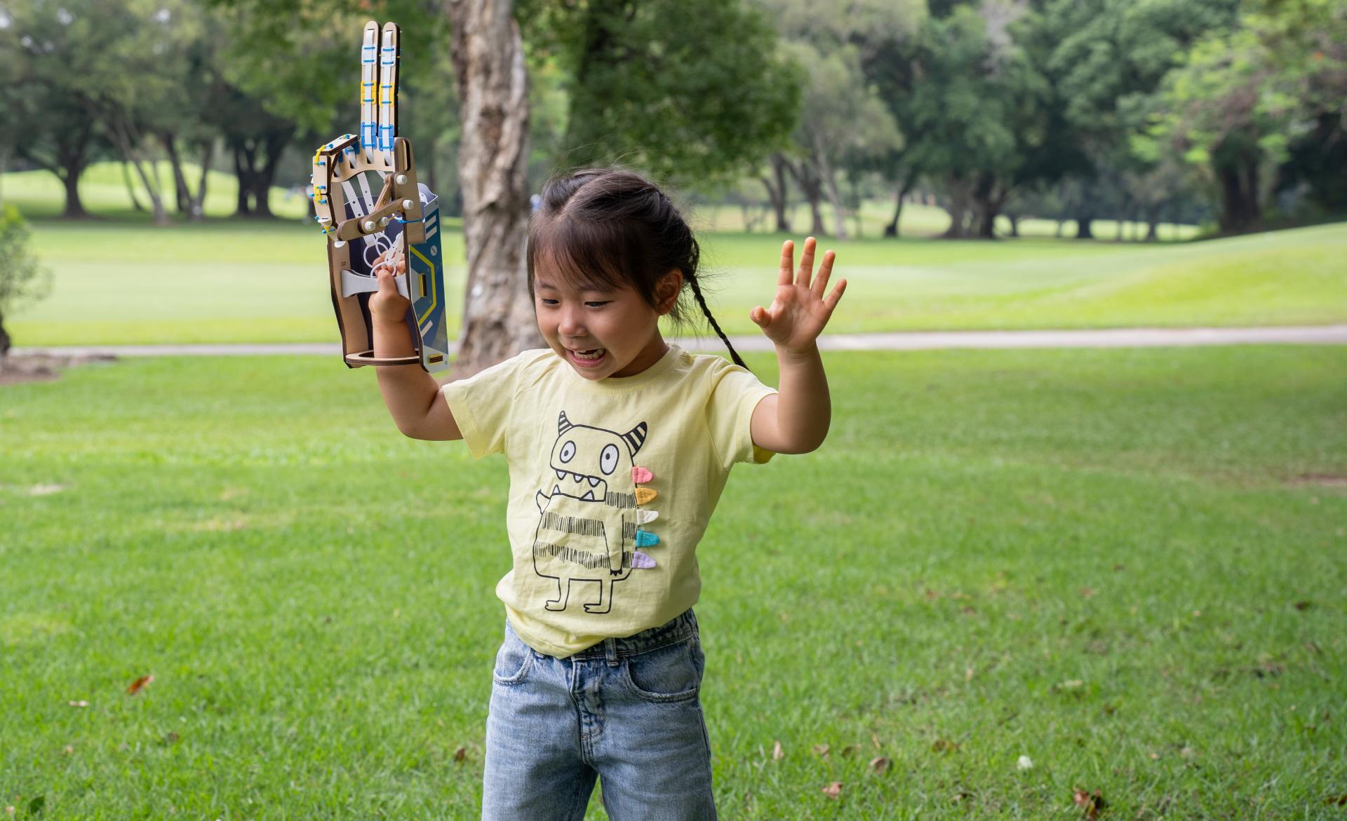 A joyful young girl with dark, braided hair laughs excitedly while playing in a grassy park. She is wearing a yellow t-shirt with a monster graphic and blue jeans. She holds up a large, wooden toy gauntlet on one arm and waves with her other hand.