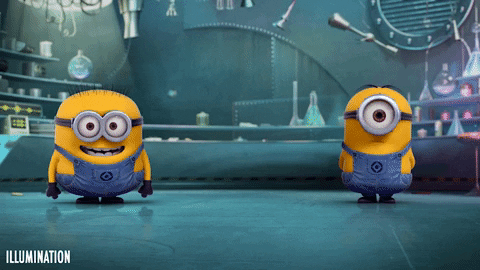 Two Minions, the small, yellow, cylindrical creatures from the Despicable Me franchise, stand on a teal floor in a laboratory-like setting. The Minion on the left has two eyes and a wide, cheerful grin. The Minion on the right has one large eye and a more neutral expression. They both wear denim overalls. The background is slightly blurred and features various laboratory equipment. The Minions shift their weight and move slightly, suggesting a playful or anticipatory mood. The Illumination logo is visible in the bottom left corner.