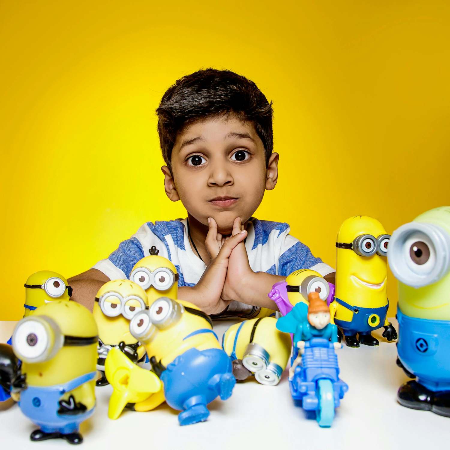 Against a bright yellow backdrop, a young boy with dark hair and a striped shirt sits at a table surrounded by a collection of Minion toys. He holds his hands together in front of his face, his expression a mix of curiosity and amusement. The Minion figures, in various poses and sizes, are scattered across the white table surface. One toy features a small figure riding a motorcycle, adding to the playful scene. The image evokes a sense of childhood fun and a fondness for the popular animated characters.