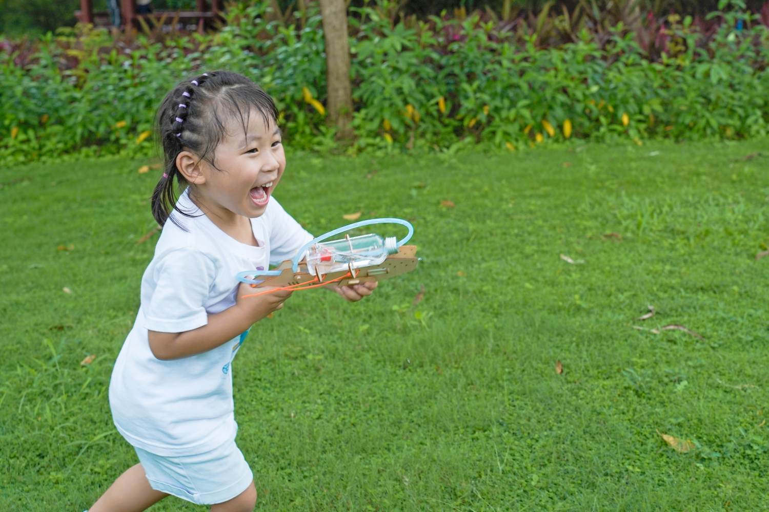 A joyful young girl with her hair in pigtails runs across a vibrant green lawn, laughing with delight as she holds a homemade-style water gun made from a plastic bottle and cardboard.