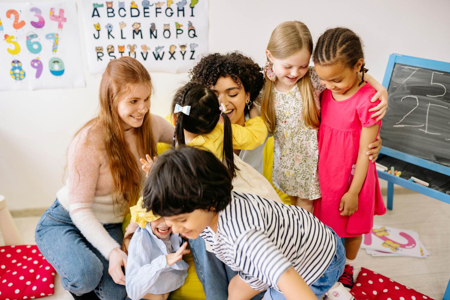 In a vibrant classroom setting, a group of preschool-aged children interacts playfully with two adults. A redhead woman sits on the floor engaging with a child, while a woman with curly hair embraces another child. Two more children stand together, arms around each other, smiling. Educational posters featuring the alphabet and numbers are visible on the wall in the background, along with a small chalkboard. The scene evokes a warm and nurturing learning environment.