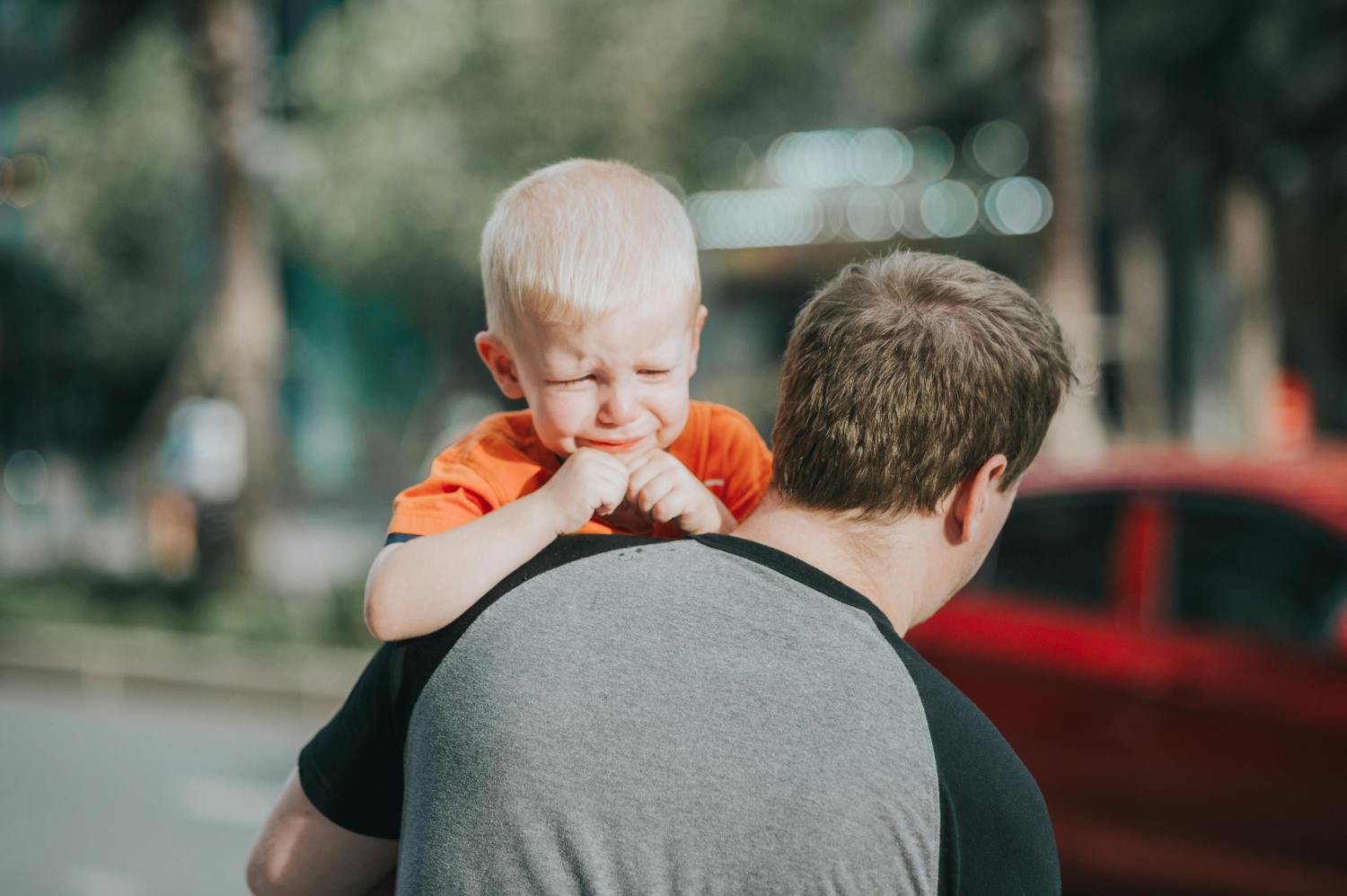 A man, seen from the back, carries a crying young boy with blond hair on his shoulders. The boy, wearing an orange t-shirt, has his eyes tightly closed and his face scrunched in distress. They are outdoors with a blurred background of trees and a red car.