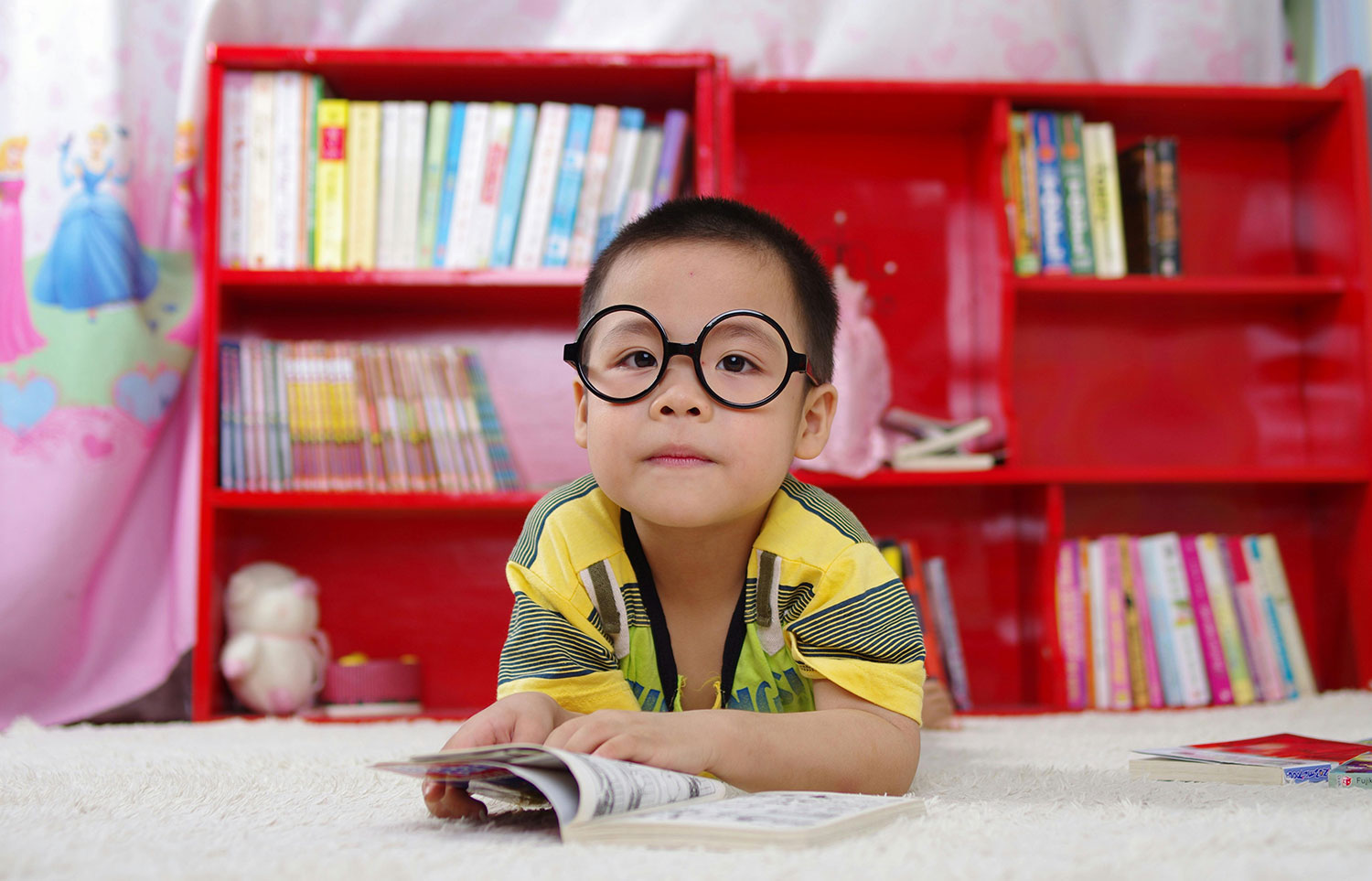 A young child wearing oversized black glasses lies on their stomach on a soft, off-white rug, engrossed in reading a comic book. They wear a yellow and green striped short-sleeved shirt. A bright red bookshelf filled with various children's books forms the backdrop, and a small stuffed animal and a pink box are visible on a lower shelf. A hint of a pink, patterned curtain or fabric can be seen to the left. The scene evokes a cozy and engaging reading environment for the child.