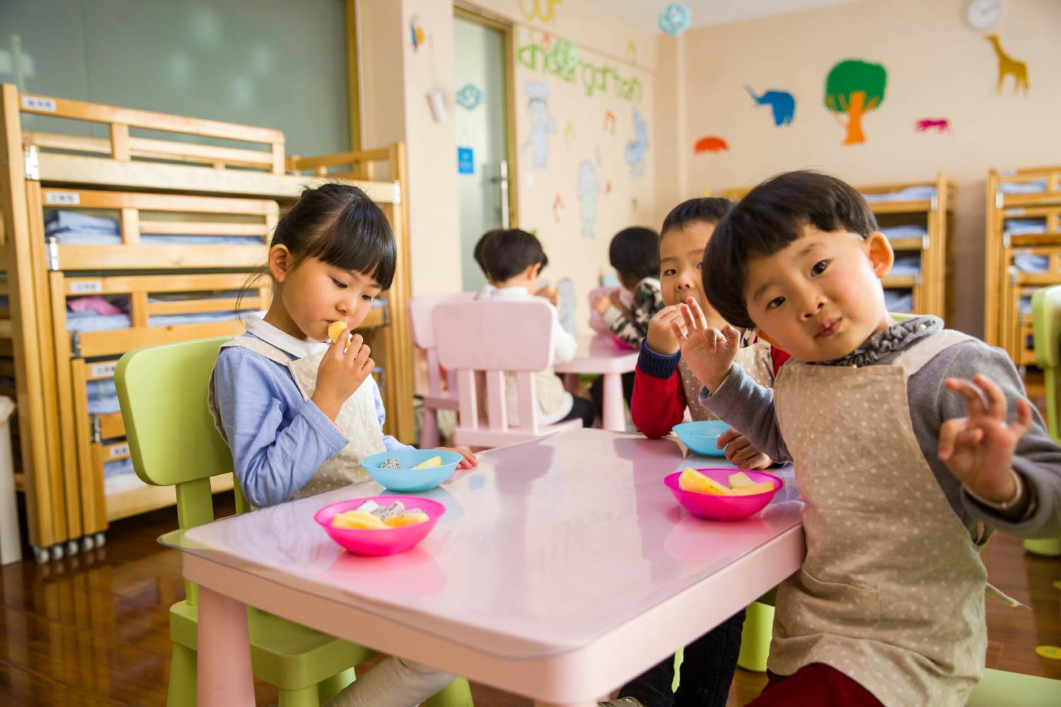 Several young children are seated at small tables in a brightly decorated preschool or kindergarten classroom. They wear aprons and are eating snacks of cut fruit from colorful bowls. One child in the foreground looks directly at the camera while gesturing with their fingers. In the background, other children are similarly engaged with their snacks. Wooden storage units are visible against the wall.