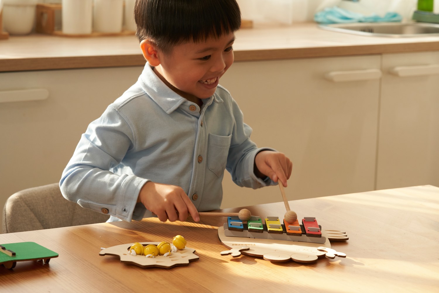 A smiling young boy in a light blue button-down shirt sits at a wooden table, happily playing a colorful, car-shaped toy xylophone with two wooden mallets. Other wooden toys are also on the table in a brightly lit room.