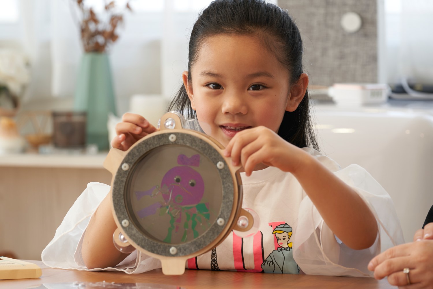A young girl with long dark hair, wearing a white t-shirt with sheer puffy sleeves, smiles at the camera as she holds up a round, wooden craft toy. The toy has a clear center displaying a purple cartoon jellyfish with green tentacles.