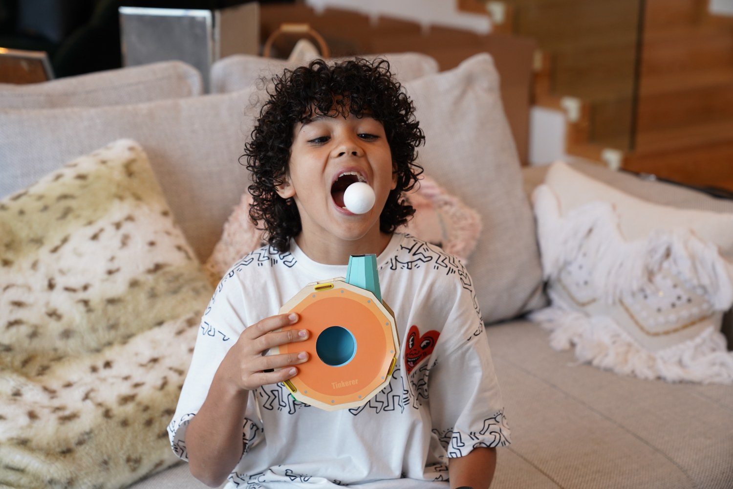 A young boy with curly hair, wearing a graphic t-shirt, sits on a couch and plays with a floating ball toy. He holds the orange and turquoise base of the toy and has a white ball in his open mouth, looking up as he plays.