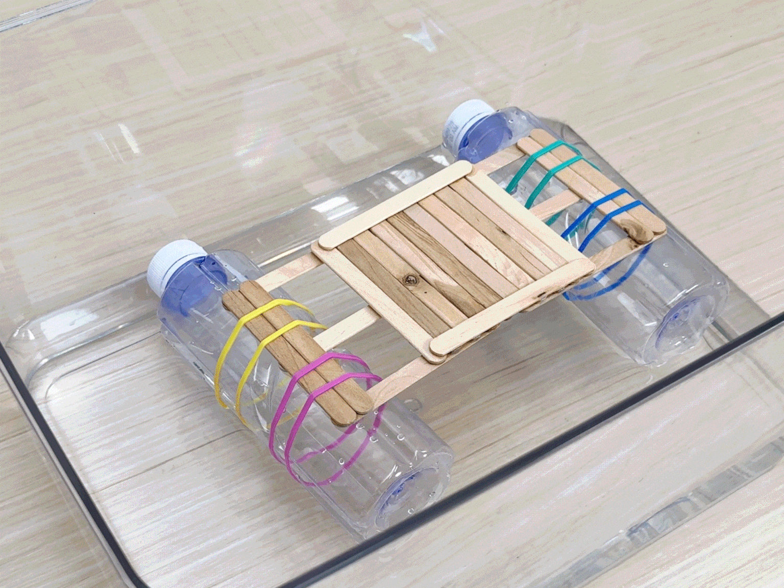 A simple raft floats in a clear plastic container filled with water. The raft is constructed from two small, clear plastic bottles secured to a platform made of flat wooden craft sticks. Colorful rubber bands hold the bottles and platform together. Two rocks are initially placed on the raft, and then a third rock is added, demonstrating the raft's buoyancy and stability. The container sits on a light wooden surface.