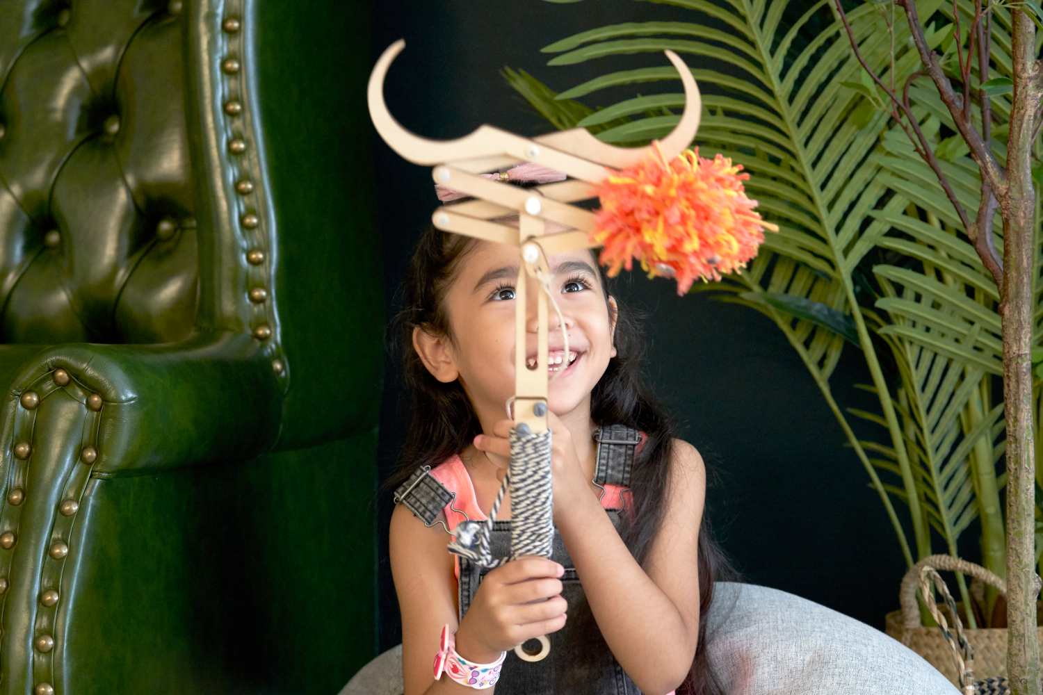 A young girl with long dark hair smiles joyfully as she looks up at a wooden toy she holds. The toy resembles a crane or grabbing device, with a bright orange and yellow pom-pom attached to its claws. She wears a denim jumper over a pink shirt and a colorful bracelet on her wrist. A dark green, tufted chair is partially visible in the background to the left, and large green plants provide a backdrop to the right. The scene has a playful and engaging atmosphere.