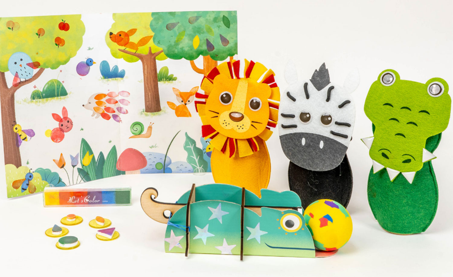 An assortment of colorful children's craft and activity items is displayed on a white background. Three felt animal figures, a lion, zebra, and crocodile, stand upright, likely designed as bowling pins. A partially assembled cardboard chameleon, a multicolored ball, small round stampers, an ink pad with a rainbow gradient, and a folded illustrated backdrop depicting a whimsical forest scene with various animals are also included. The arrangement suggests a playful and creative activity kit for children with an animal or jungle theme.