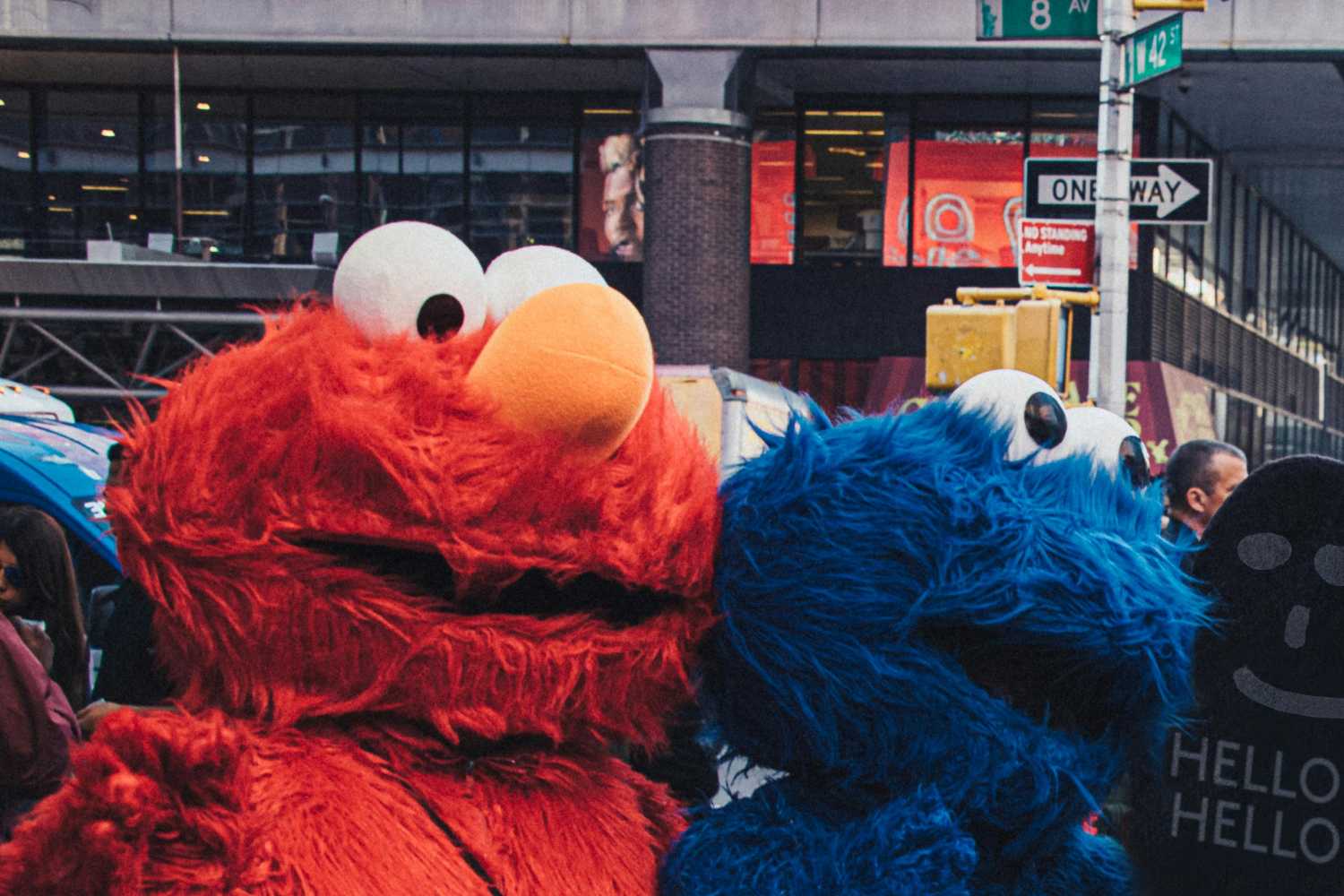 Elmo and Cookie Monster, two beloved characters from Sesame Street, are seen in a close-up shot, likely on a city street. Elmo, a vibrant red, furry Muppet with large white eyes and an orange nose, dominates the left side of the frame. Cookie Monster, a bright blue, furry Muppet with similar large white eyes, fills the right side. The background, slightly blurred, suggests a busy urban environment with buildings and street signs visible. A partially visible sign reading HELLO HELLO is held up near Cookie Monster.