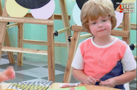 A young child with light, short hair and a peach and blue t-shirt sits at a table. They are focused on manipulating a small object in their hands, possibly playdough or clay. In the background, wooden easels with circular color palettes are visible. The text The Secret Life of 4 Year Olds appears in the upper right corner. This video likely shows a child engaged in a creative activity in a preschool or playroom setting.