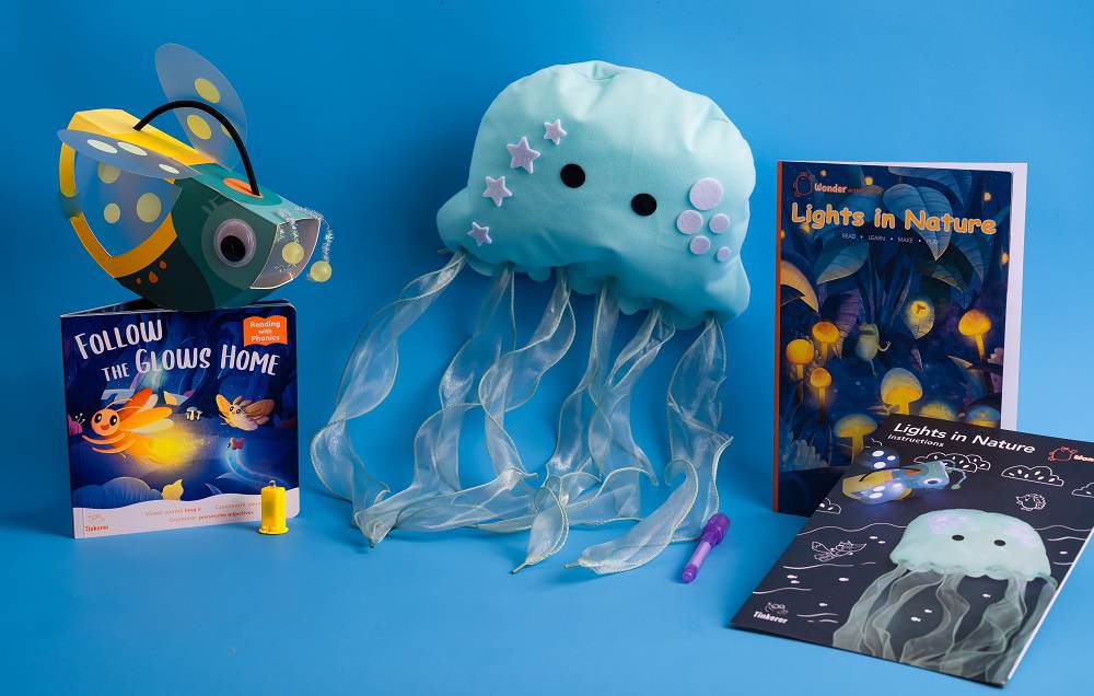 A collection of children's educational materials themed around bioluminescence is displayed on a vibrant blue background. A plush light blue jellyfish toy with sheer, iridescent tentacles is positioned centrally. Beside it are a booklet titled Lights in Nature with a cover depicting glowing fungi and a smaller instruction booklet for a related activity. A children's book, Follow the Glows Home, featuring a firefly, and a constructed paper firefly lantern complete the arrangement. A small yellow marker lies near the jellyfish.