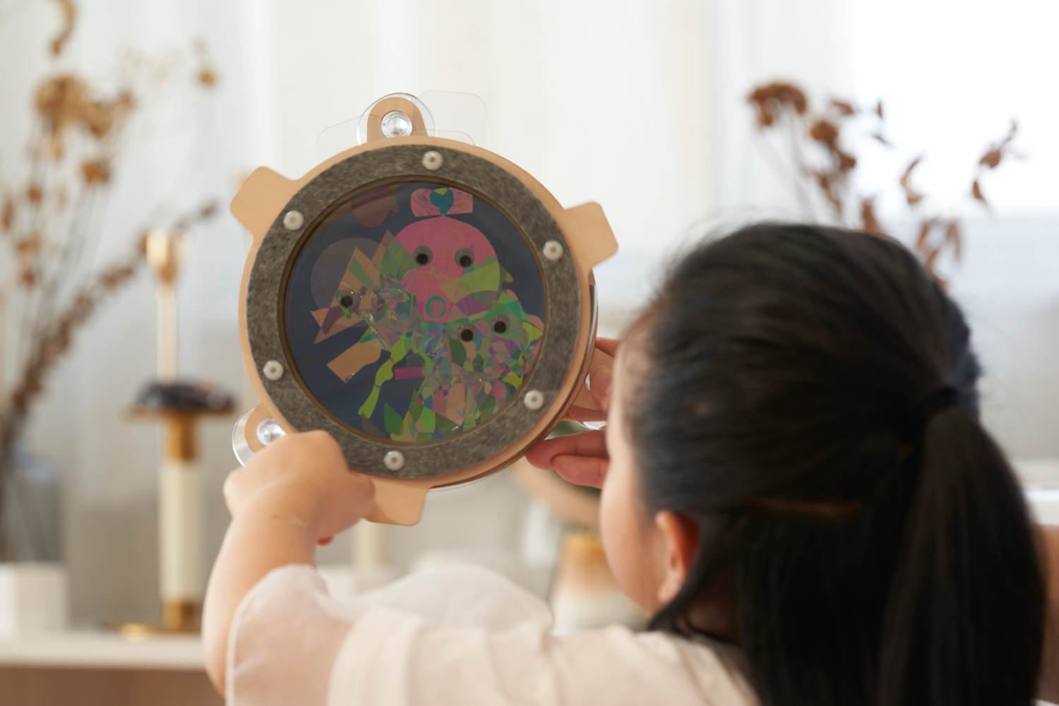 A young child with dark hair pulled back in a ponytail holds a circular wooden toy with a clear center filled with colorful, translucent shapes. The child, seen from over their shoulder, is focused on manipulating the toy, which appears to be designed for visual stimulation and play. The background is softly blurred, showing a light-colored interior space with hints of dried flowers or plants.