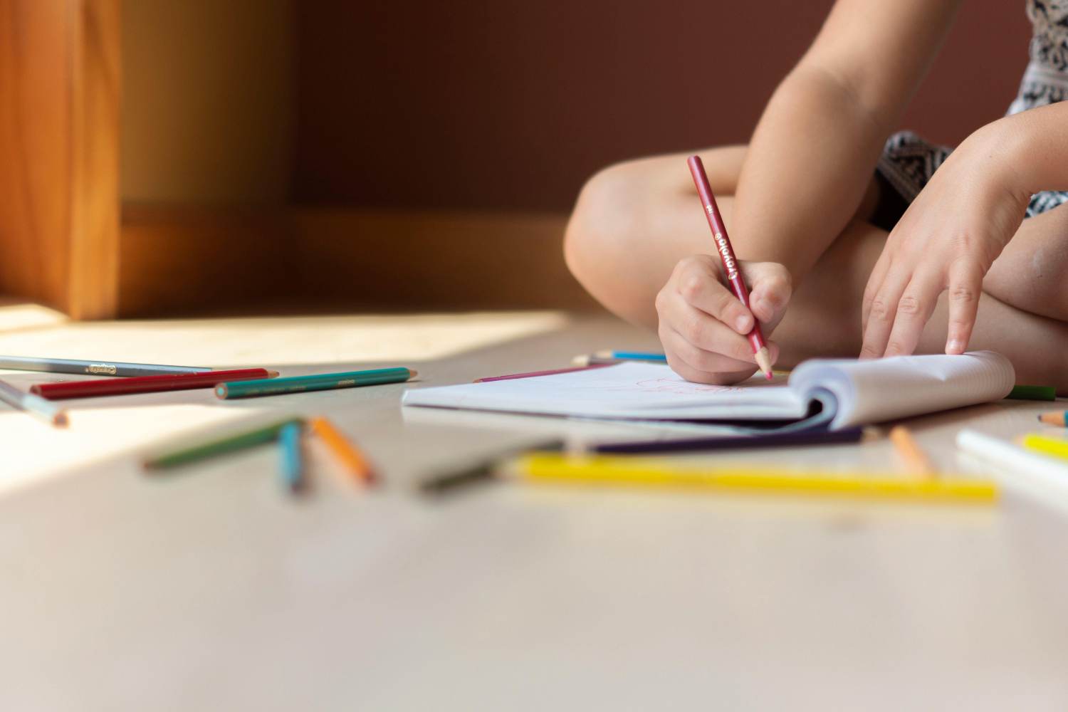 A child sits on a light-colored floor, legs tucked underneath, drawing in a sketchbook with a red colored pencil. Their left hand rests on the page while their right hand carefully maneuvers the pencil. Several other colored pencils are scattered around the sketchbook on the floor. A portion of a wooden piece of furniture is visible in the background, blurred and out of focus. Sunlight streams in from the left side of the image, illuminating the scene.