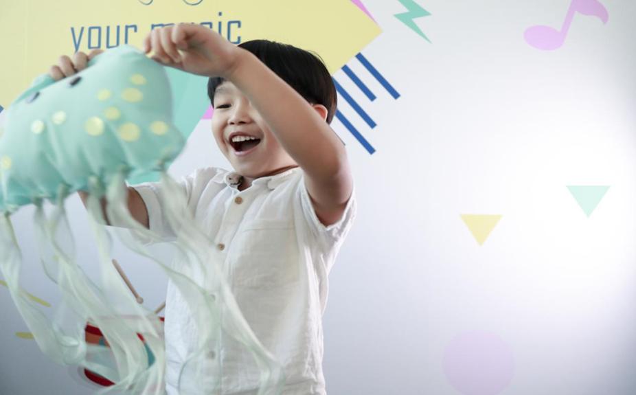 The image shows a young boy, happily holding a light teal jellyfish-shaped toy. The toy has yellow polka dots and long, flowing tentacles. The boy is wearing a white, button-down shirt and is smiling broadly. He appears to be indoors, with a brightly colored wall in the background featuring musical notes and geometric shapes. The overall mood of the image is cheerful and playful.