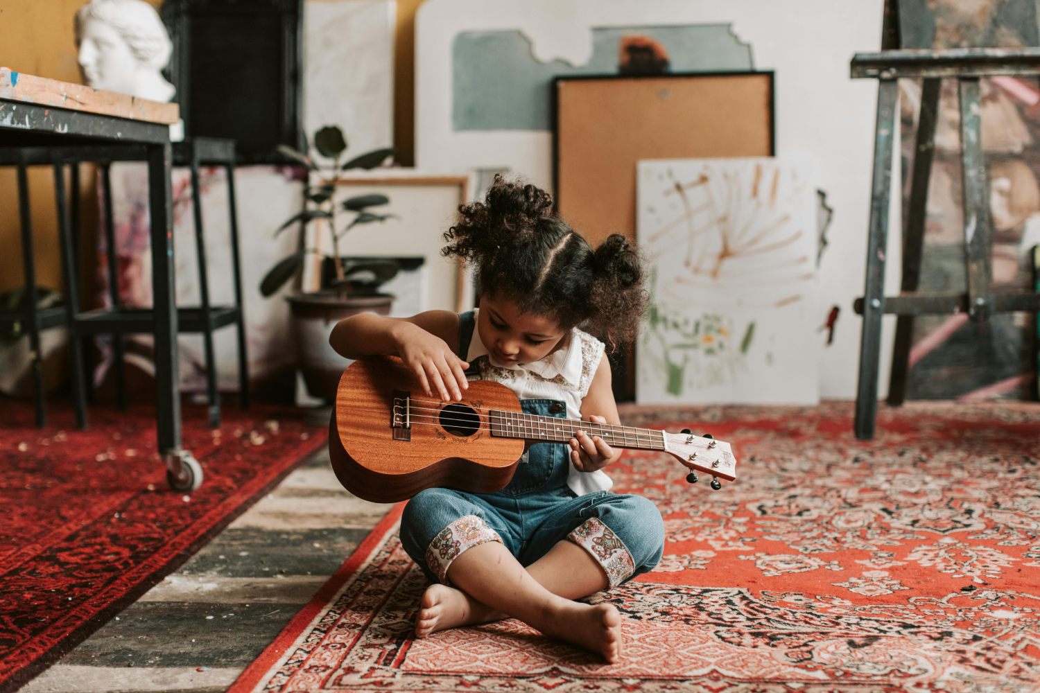 A young girl with curly hair in two puffs sits cross-legged on a red patterned rug in an artist's studio, looking down as she plays a small wooden ukulele. The room around her is filled with art supplies, including canvases, an easel, and a sculpture.