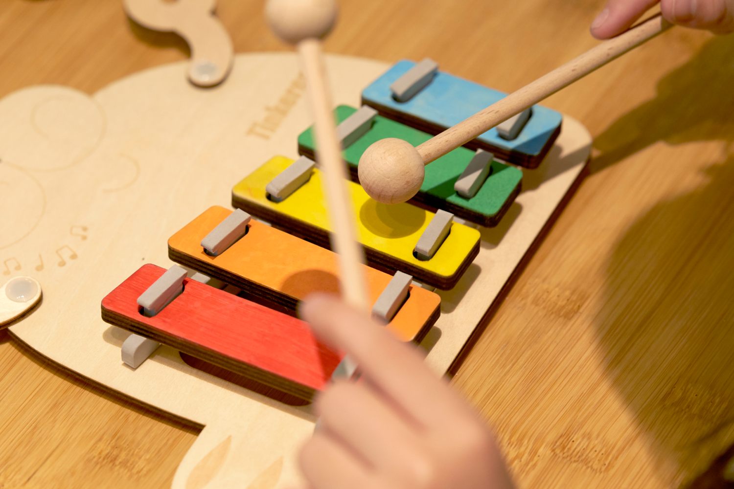 A close-up of a person playing a colorful wooden toy xylophone with two mallets. The instrument has red, orange, yellow, green, and blue keys on a light wood base, all resting on a wooden table. One of the mallets is in motion, blurring as it's about to strike a key.