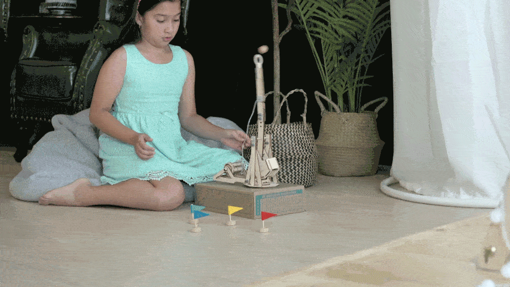 A young girl, comfortably seated on the floor, prepares to launch a small wooden ball using a miniature catapult. This STEM-oriented toy, crafted from wood, demonstrates basic physics principles like trajectory and force as the girl aims for colourful flags positioned a short distance away.